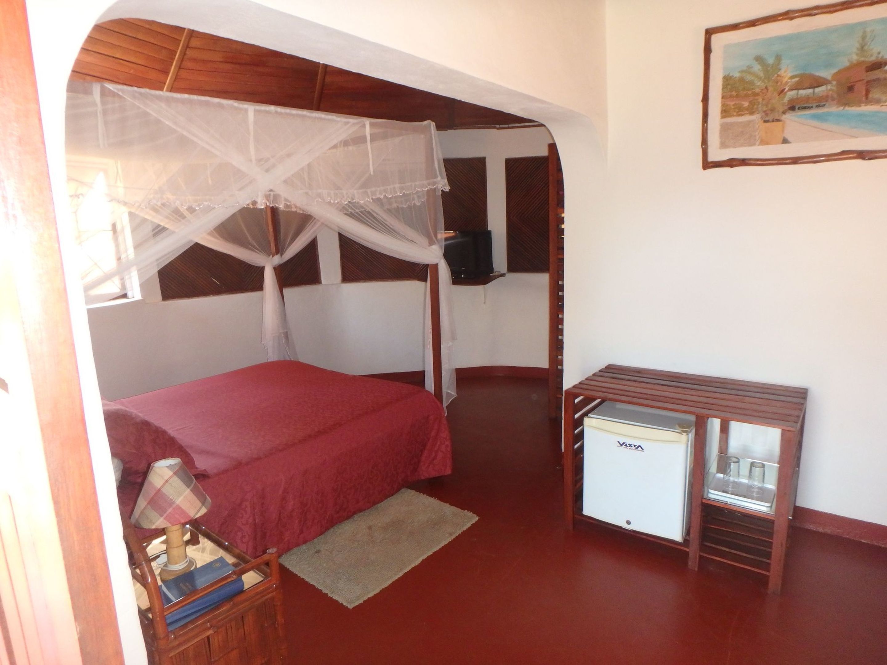 A bedroom with a bed under a mosquito net, a small table with a refrigerator, and a painting on the wall.