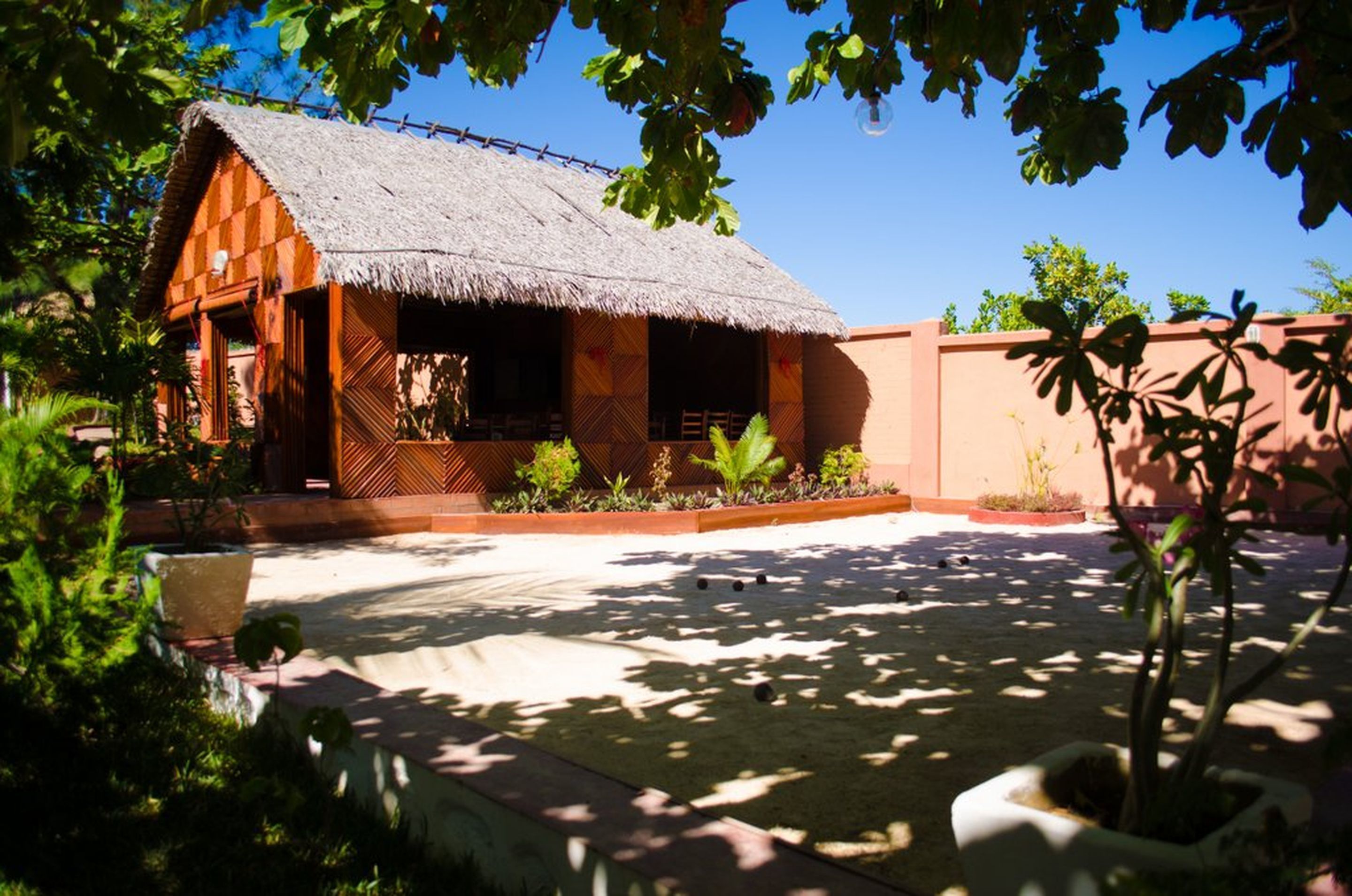 Un bungalow con techo de paja y un pequeño porche, rodeado de exuberante follaje y un patio empedrado.