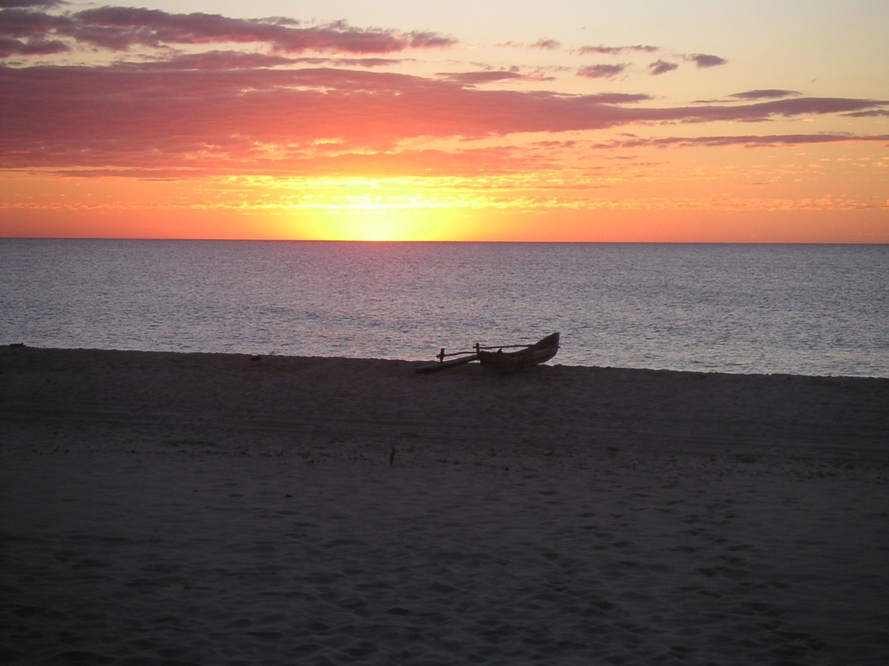 Cena de praia com um barco na areia, perto da margem da água durante o pôr do sol.