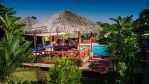 A thatched-roof outdoor seating area with a pool, surrounded by lush greenery. - Edena Kely (Mahajanga)