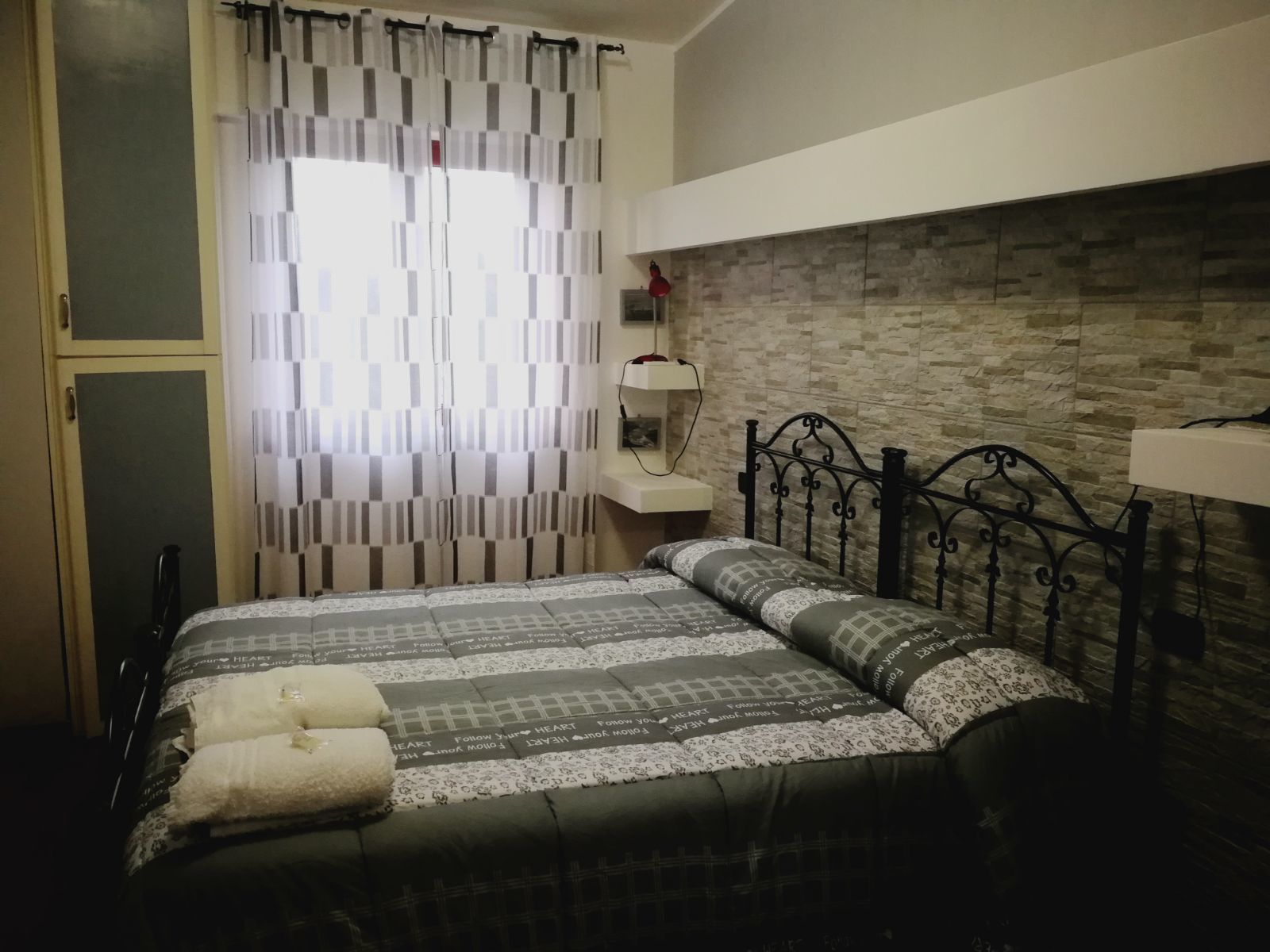 A bedroom with a bed, a headboard, a bedside table, a mirror, and a window with curtains.