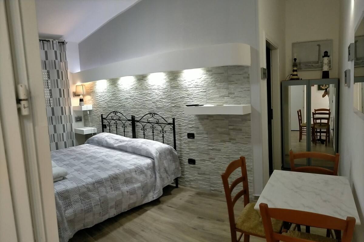 Un dormitorio con cama, mesita de noche, lámpara, espejo y un área de comedor con mesa y sillas.