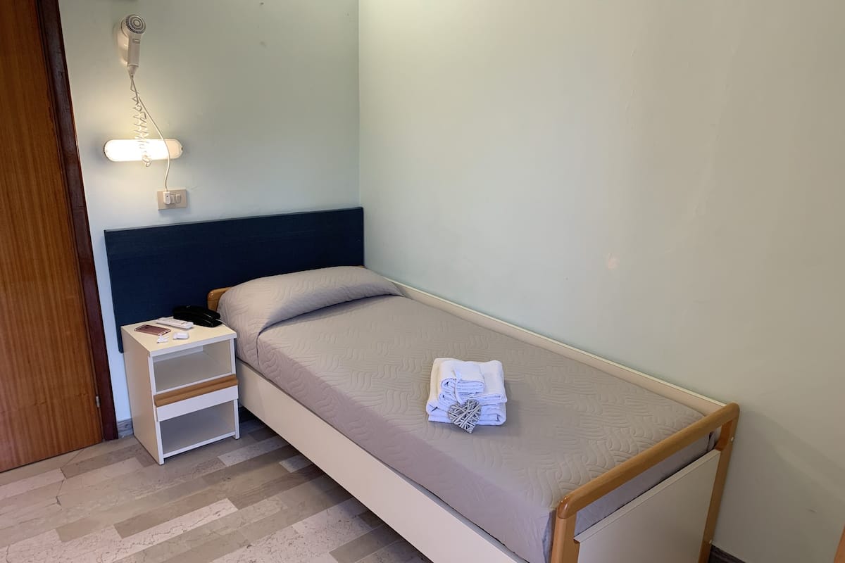 Quarto pequeno com uma cama de solteiro, cabeceira de madeira, mesa de cabeceira com abajur e uma toalha dobrada na cama.