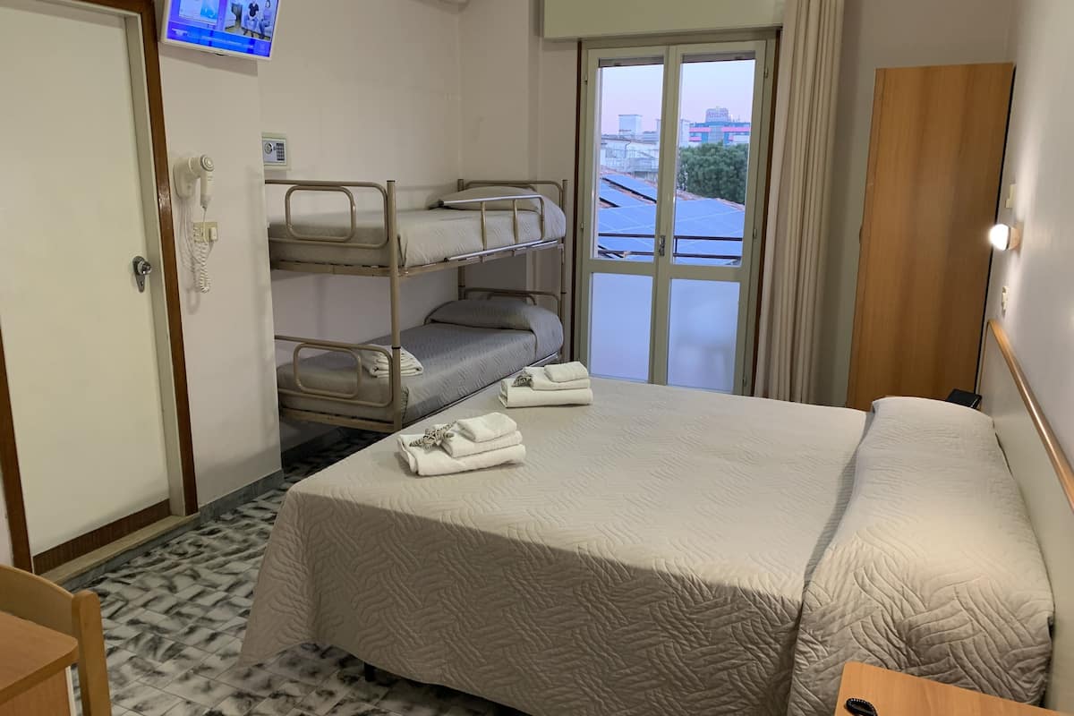Quarto de hotel com cama beliche, uma cama de solteiro, televisão montada na parede e janela com vista para edifícios.