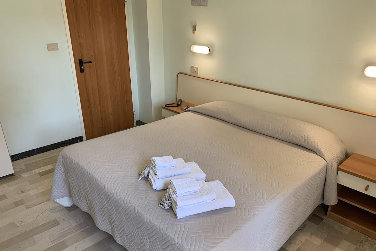 Cama bem arrumada com colcha bege e toalhas dobradas, porta de madeira, mesa de cabeceira com abajur e luminária na parede.