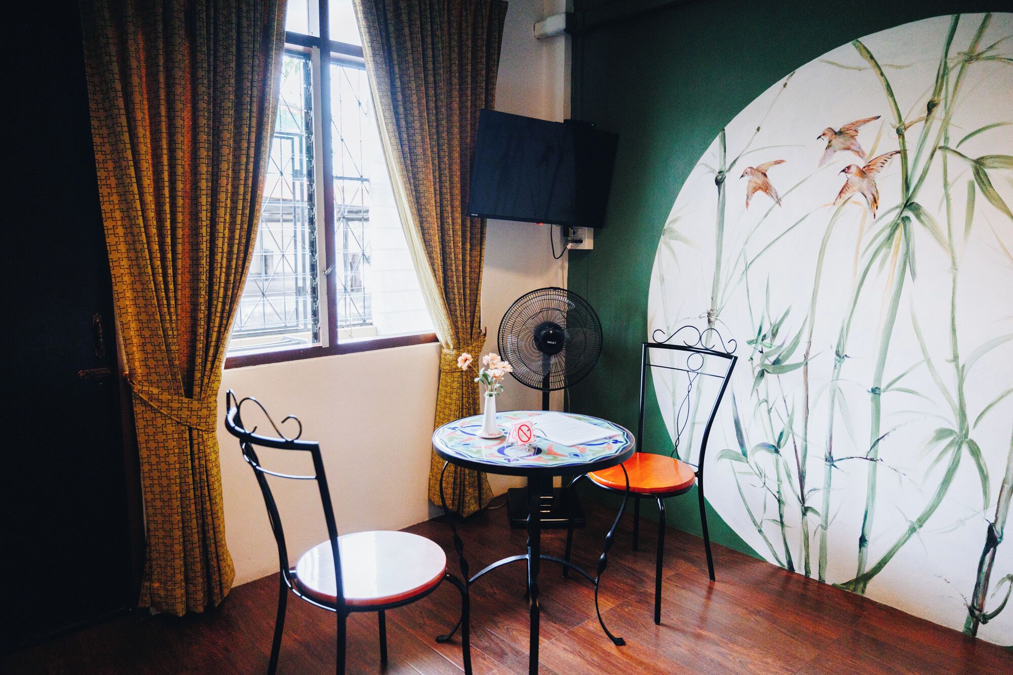A room with a table and chairs, a window with curtains, a wall-mounted fan, and a large wall mural.