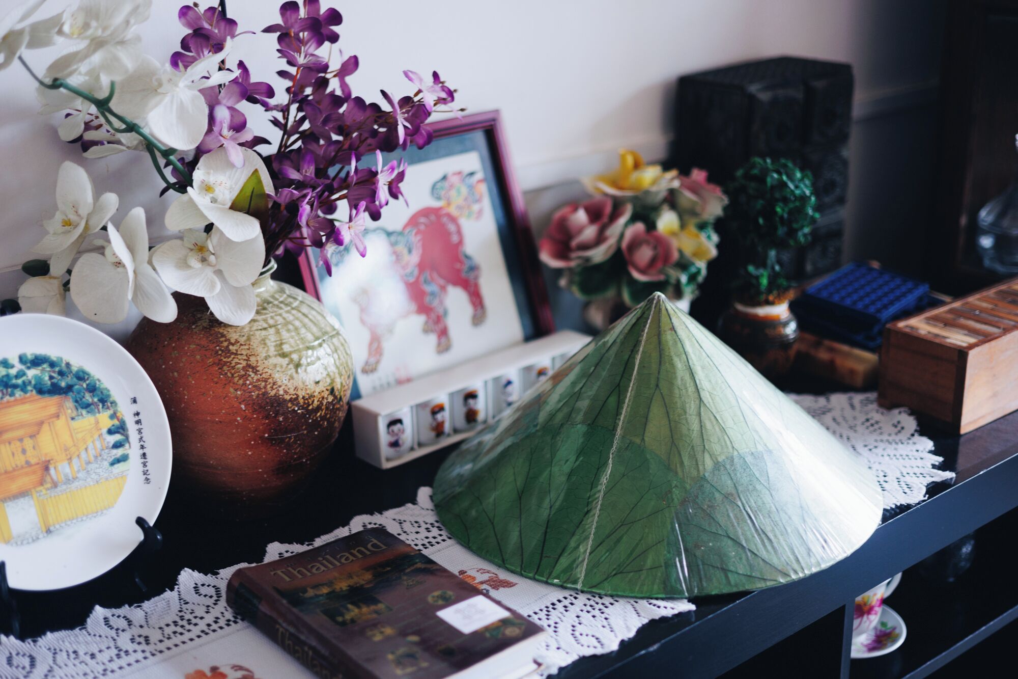 A table with a green leaf, a book titled 'Thailand', a vase with white and purple flowers, and a framed picture.