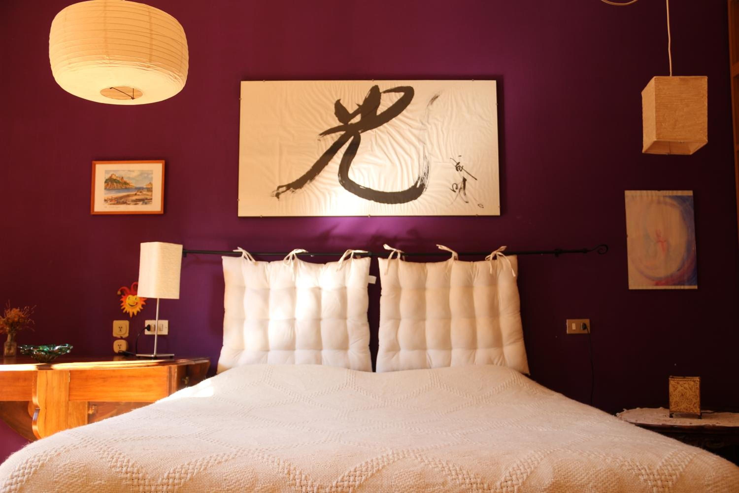 Una cama con ropa de cama blanca, una gran obra de caligrafía enmarcada en la pared y una lámpara colgante.