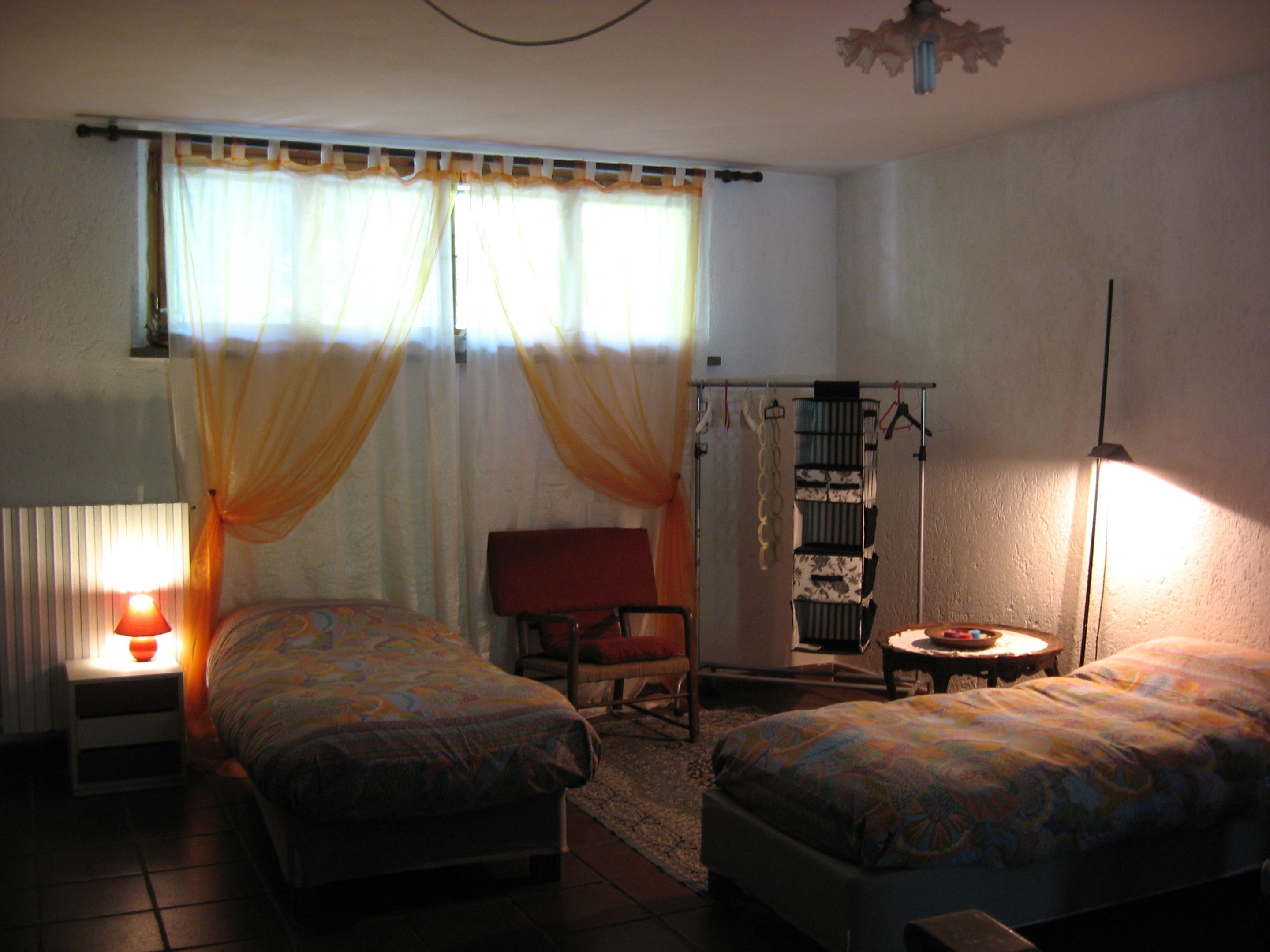 Una camera da letto con due letti, una sedia, un tavolino, un termosifone e una finestra con tende.