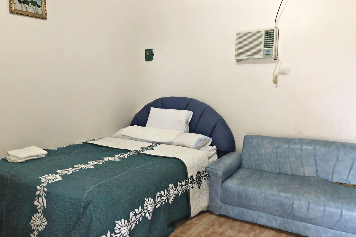 A bedroom with a bed, a blue sofa, a wall-mounted air conditioner, and a small green device on the wall.