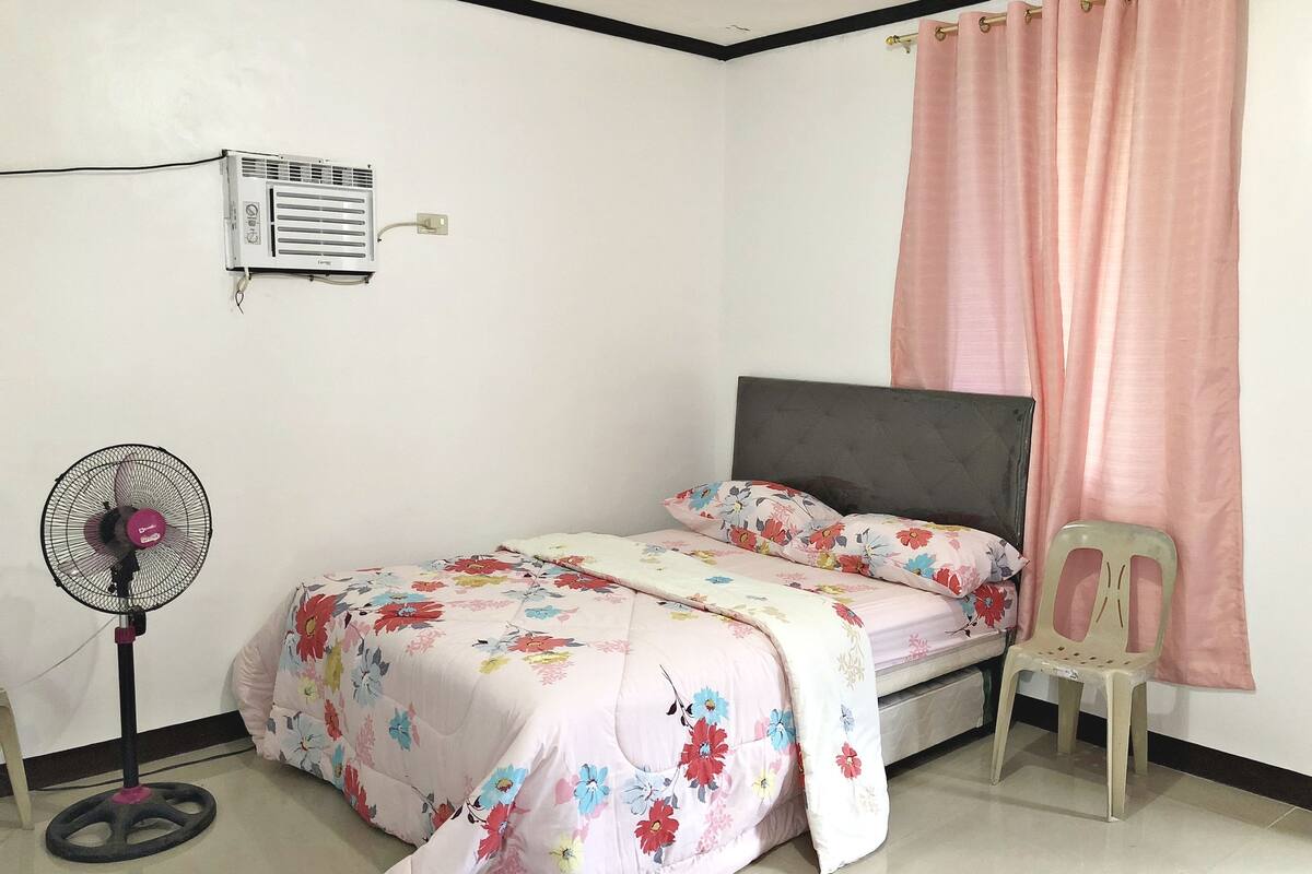 A bedroom with a bed, a standing fan, a plastic chair, and a window with a pink curtain.