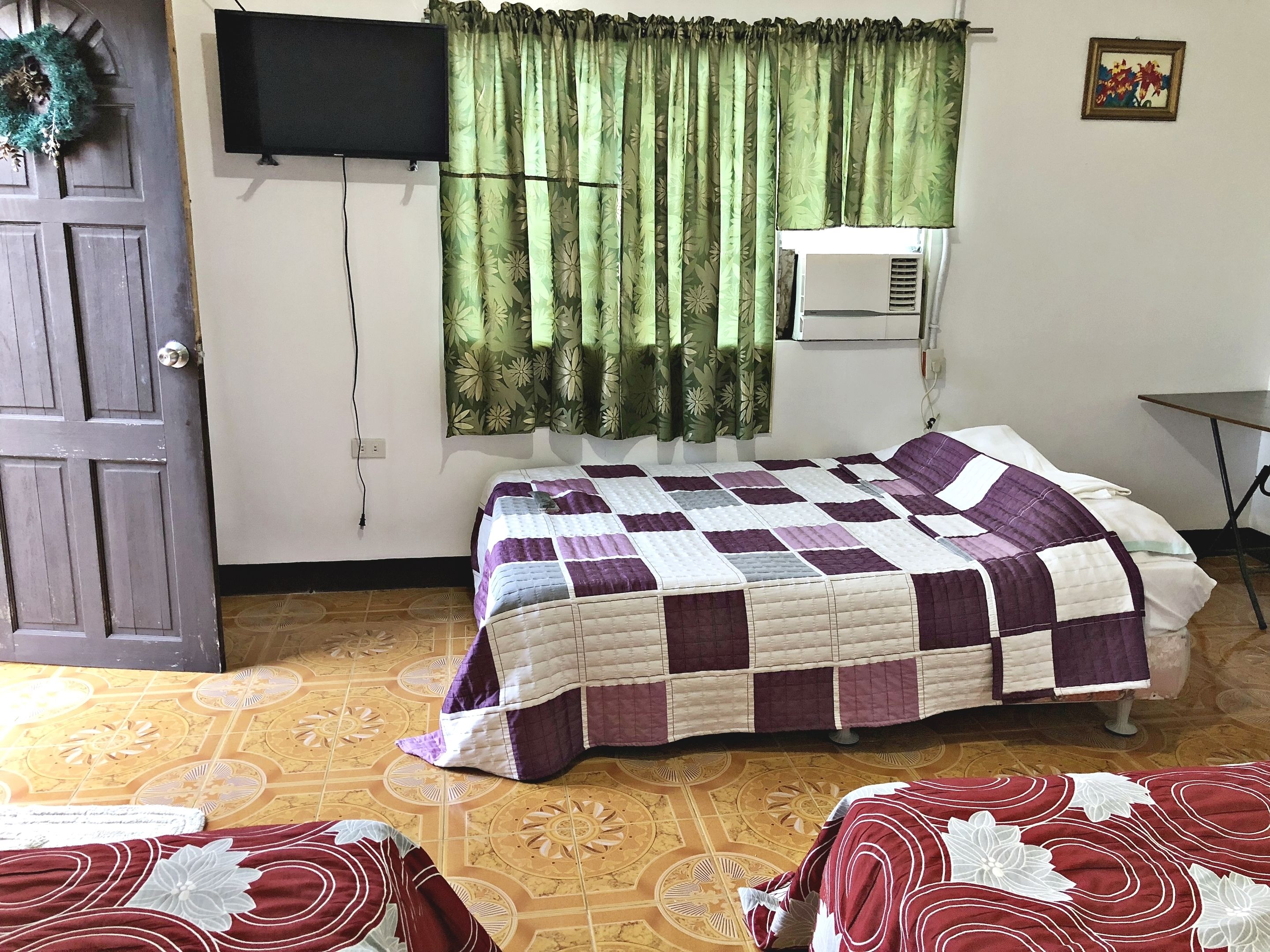 Una habitación con una cama, un televisor en la pared, una ventana con cortinas verdes y una mesita.