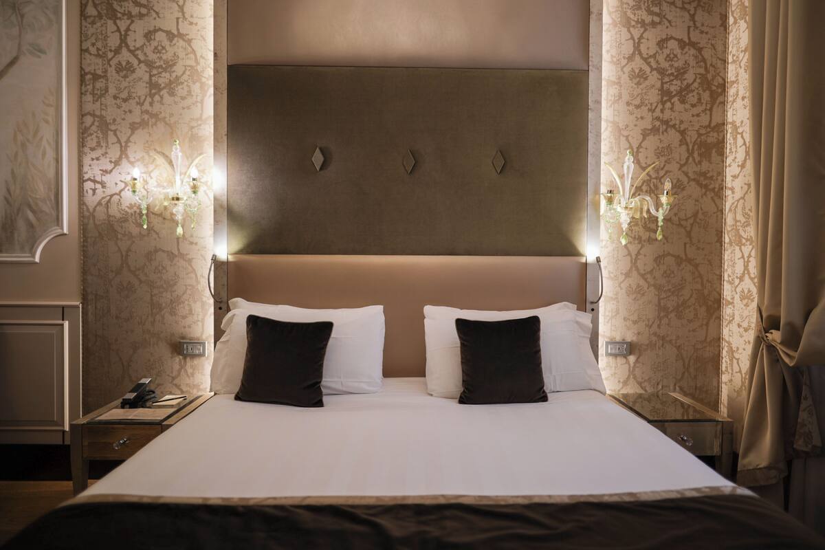Une chambre d’hôtel avec un grand lit, deux tables de chevet, une petite table et une tête de lit agrémentée d’appliques murales.