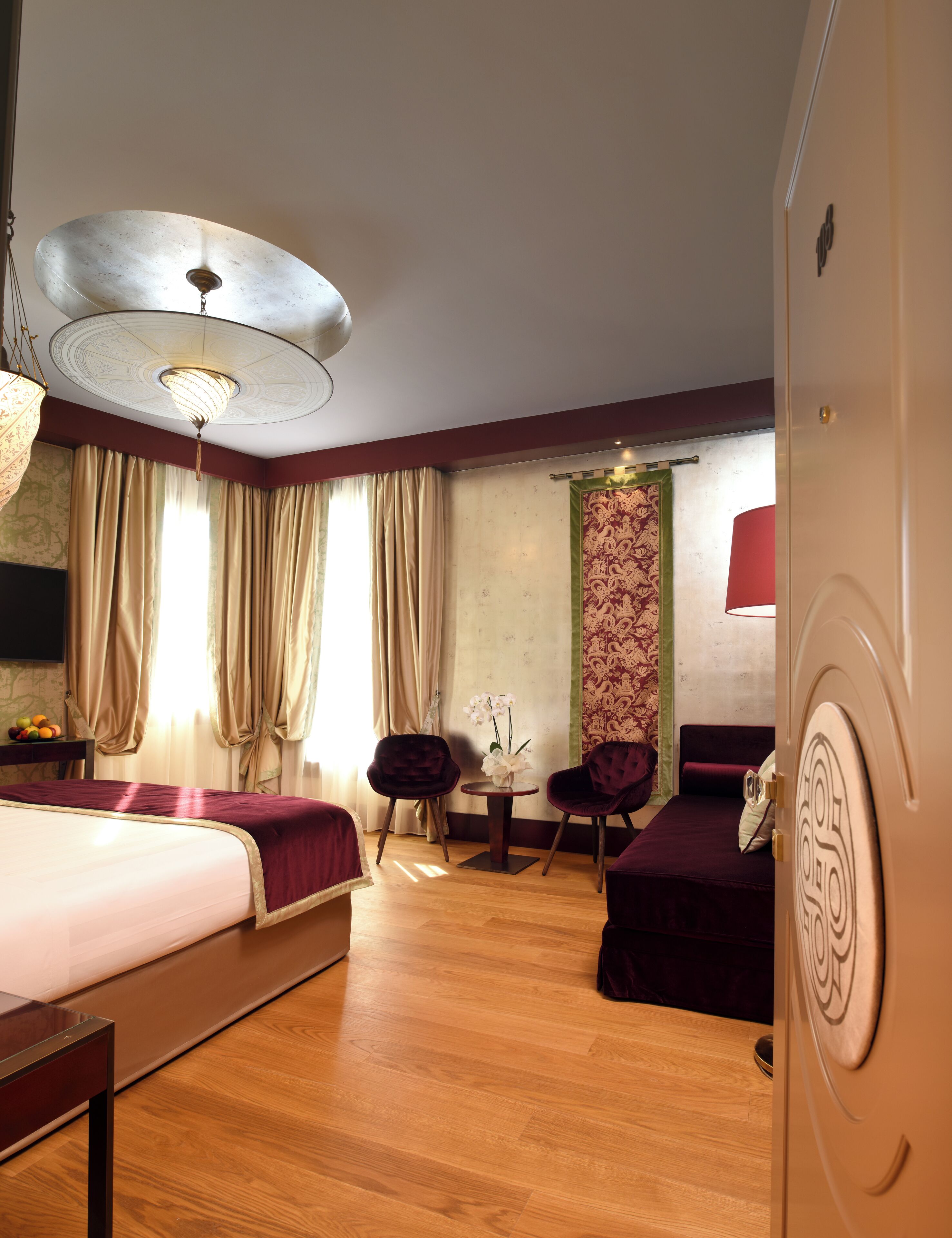 Camera d'albergo con un letto, due poltrone, un tavolino con fiori, una lampada rossa e un ventilatore a soffitto.