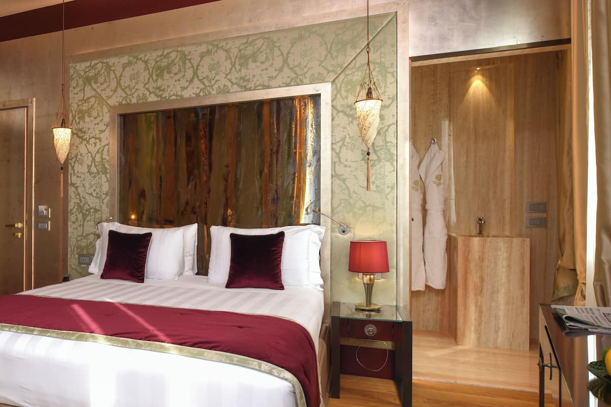 Une chambre d’hôtel avec un grand lit, un coin salon avec un fauteuil, une table de chevet avec une lampe, et une salle de bain visible à travers une porte ouverte.