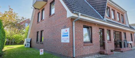 Ein zweistöckiges Backsteingebäude mit geneigtem Dach und einem Schild, das es als Mehrfamilienhaus kennzeichnet. Vor dem Haus befindet sich ein kleiner Garten mit Rasenfläche und Baum.