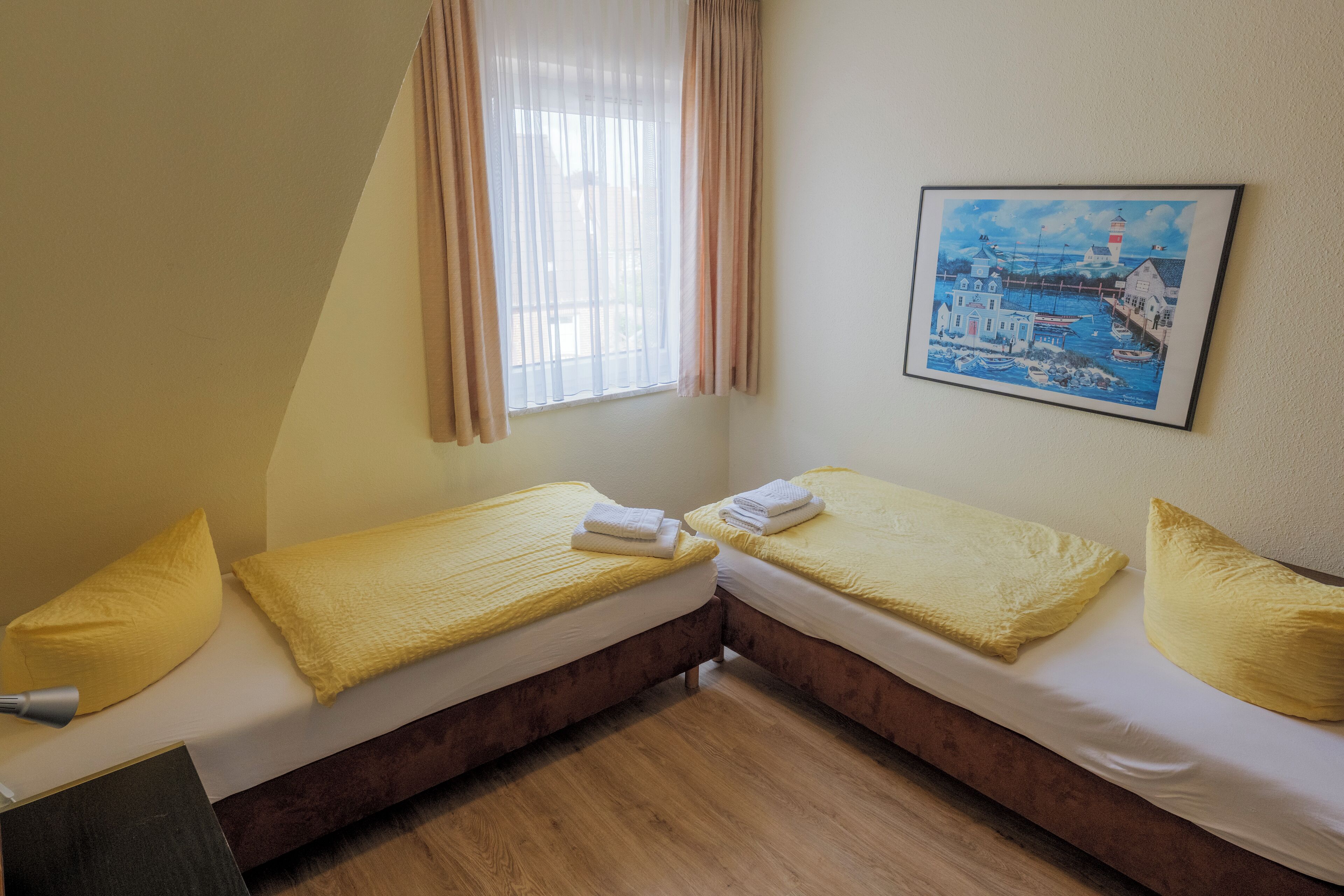 A room with two beds, each with a yellow blanket and pillow, a painting of a coastal scene, and a window with sheer curtains.