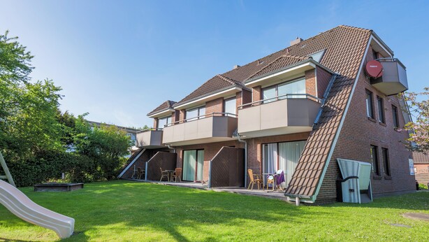 A two-story house with a sloped roof, a slide in the yard, and a small patio with chairs and a table. - Appartementhaus Alt B üsum (Büsum)