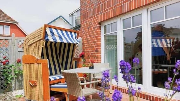 A wicker outdoor seating area with a table and chairs, a brick house with a large window, and purple flowers in the foreground. - Boardinghouse Büsum (Büsum)