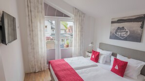 A hotel room with a bed, a window with curtains, a painting on the wall, and a view of buildings outside. - Boardinghouse Büsum (Büsum)