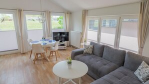 A living room with a grey sofa, a white coffee table, and a TV. There is a dining table with chairs and a small potted plant. - Boardinghouse Büsum (Büsum)