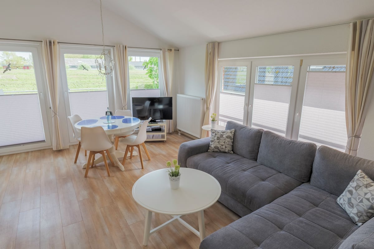 Una sala de estar con un sofá gris, una mesa de centro blanca y un televisor. Hay una mesa de comedor con sillas y una pequeña planta en maceta.