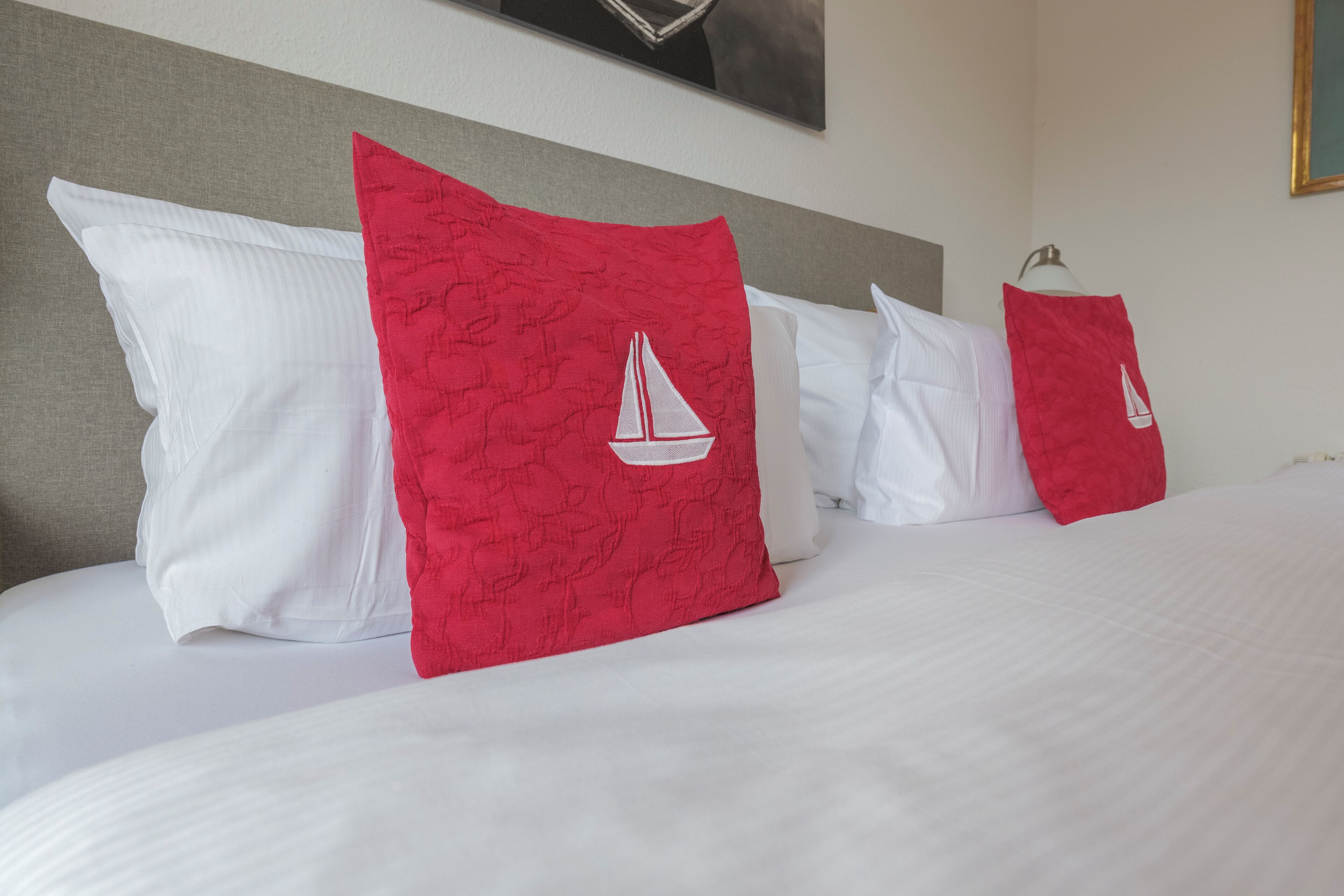 A neatly made bed with white pillows and red pillows featuring a sailboat design.