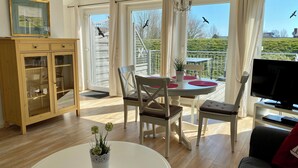 A spacious living area with a dining table, chairs, a TV, and a view of a balcony with a railing and greenery. - Boardinghouse Büsum (Büsum)
