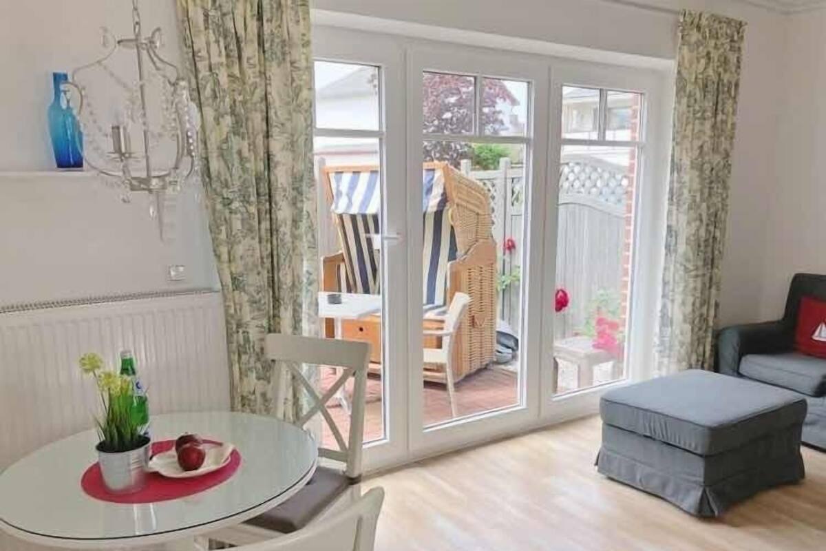 Una habitación con una mesa de vidrio, sillas blancas, un sofá gris y un ventanal corredero que da a un espacio exterior con una silla de mimbre y una silla a rayas.