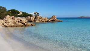 Ein Strand mit klarem türkisblauem Wasser, Felsformationen und ein Gebäude mit einem Baum auf der linken Seite.