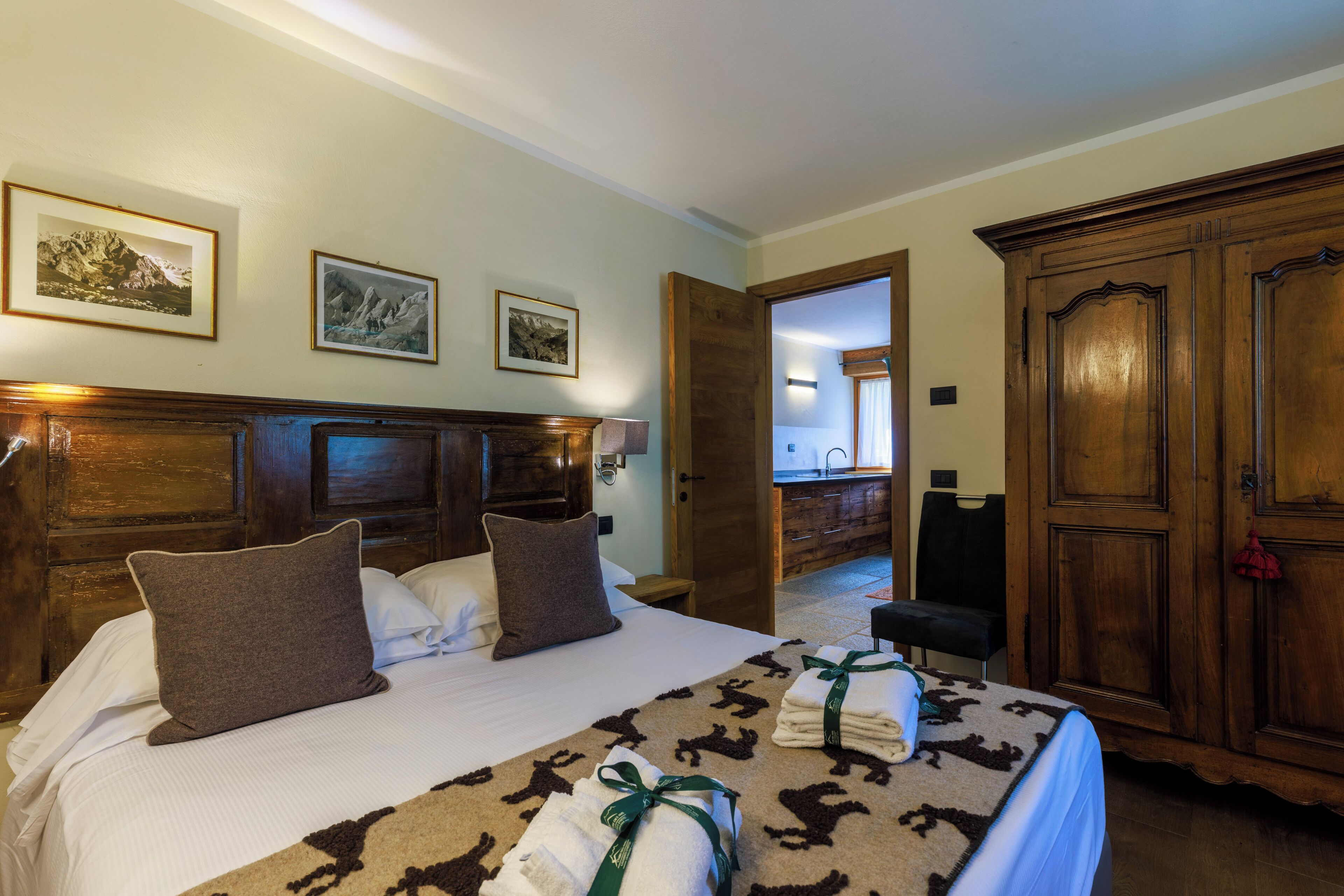 A hotel room with a large bed, wooden headboard, and a wardrobe. There are framed pictures on the wall and a bathroom visible through an open door.