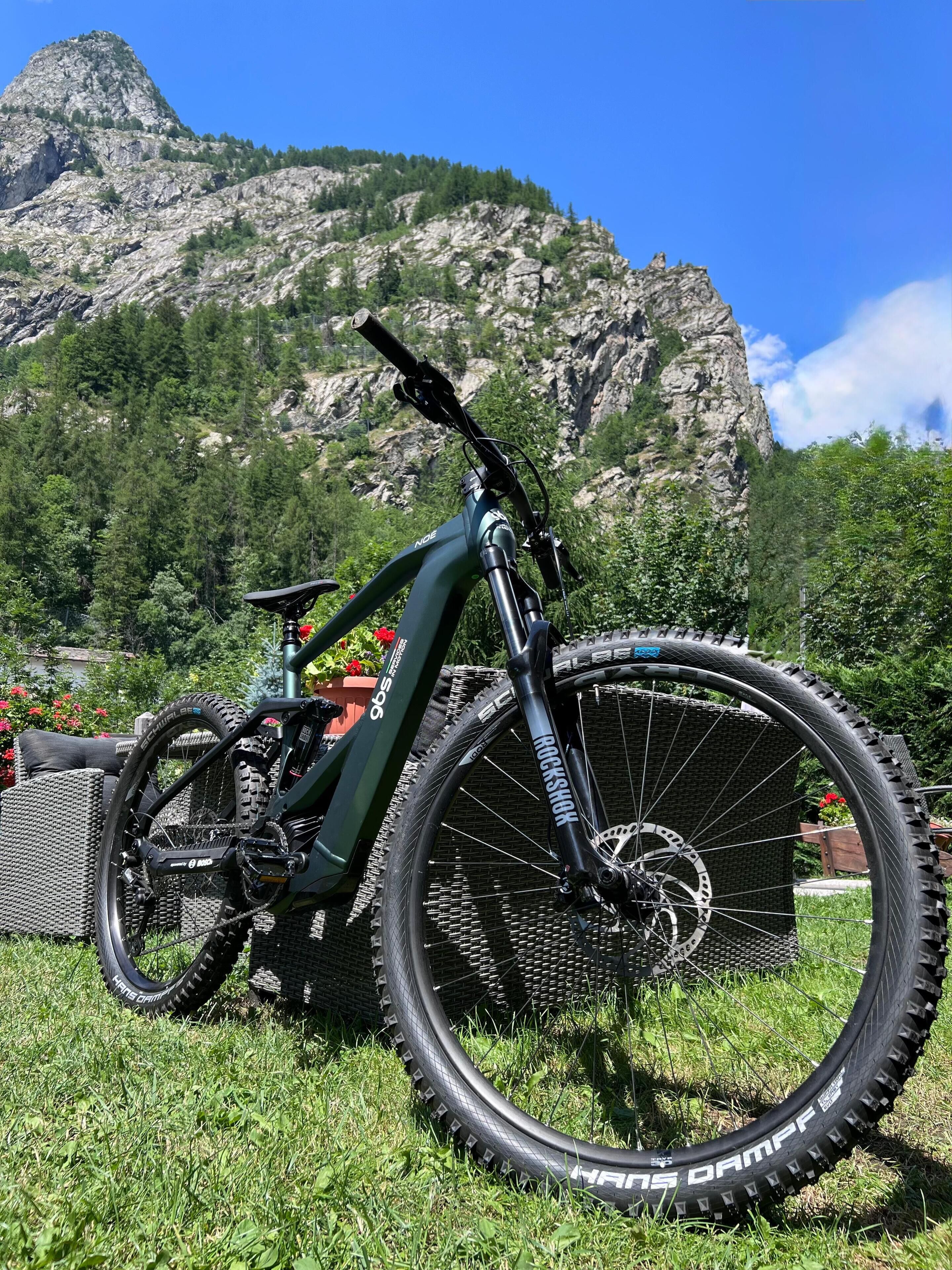 Una bicicleta de montaña con suspensión delantera, un cambio trasero y una cadena, sobre césped, con una silla de mimbre y una planta en maceta en primer plano, y una montaña rocosa al fondo.