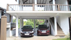 A covered car parking area with two cars parked, a staircase to the right, and a white building with columns. - Selfcate Luxury Apartments (Kandy)