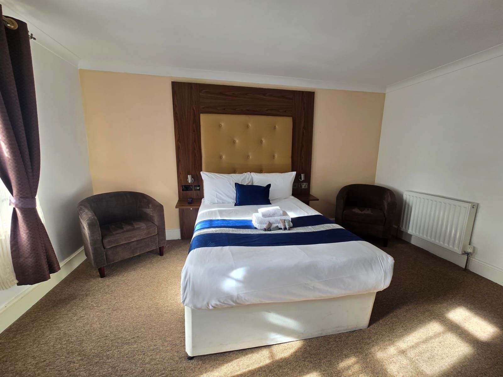 A hotel room with a large bed, a wooden headboard, a nightstand, a chair, and a radiator.