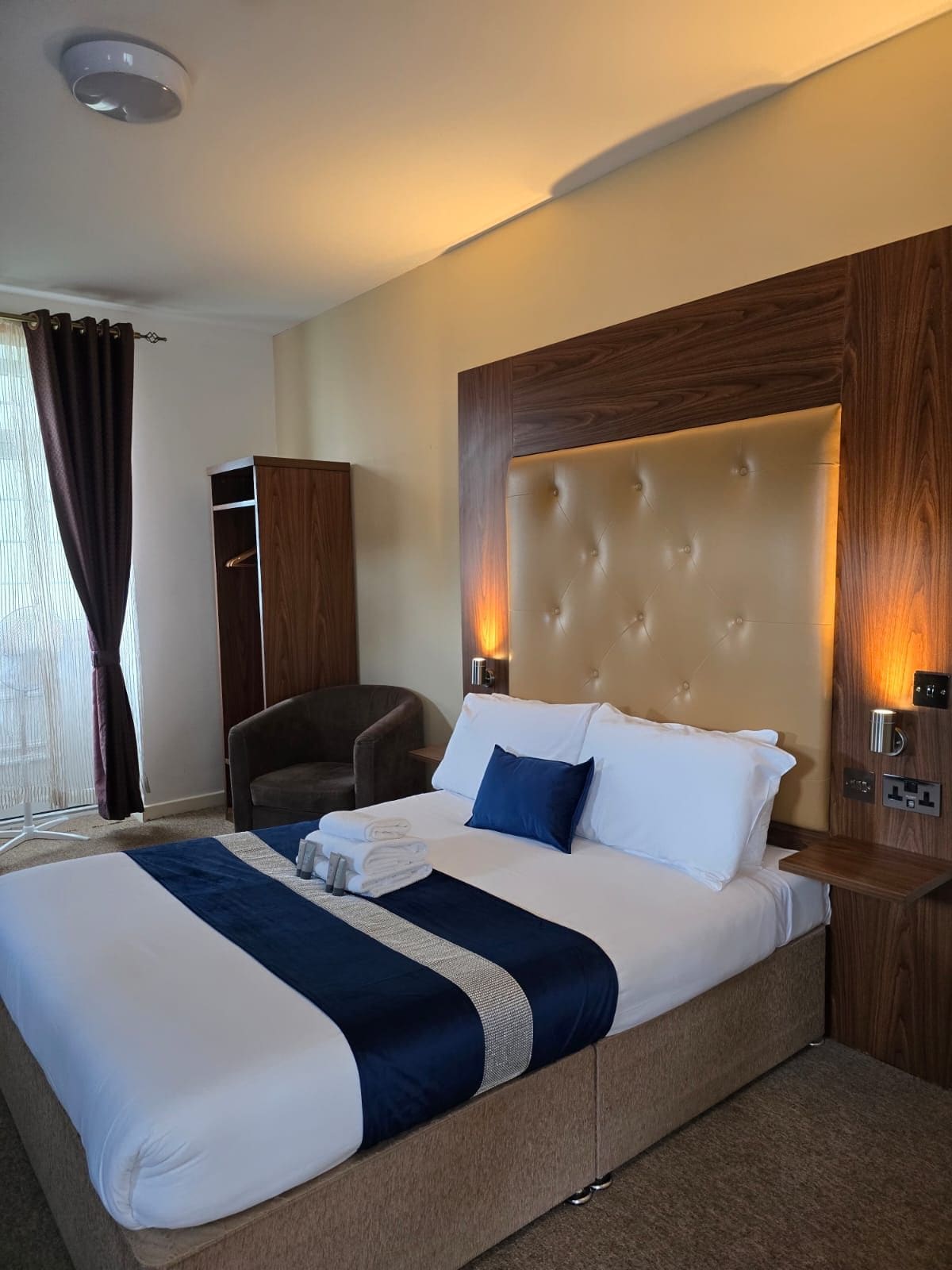 A hotel room with a large bed, a wardrobe, a chair, and a bedside table.