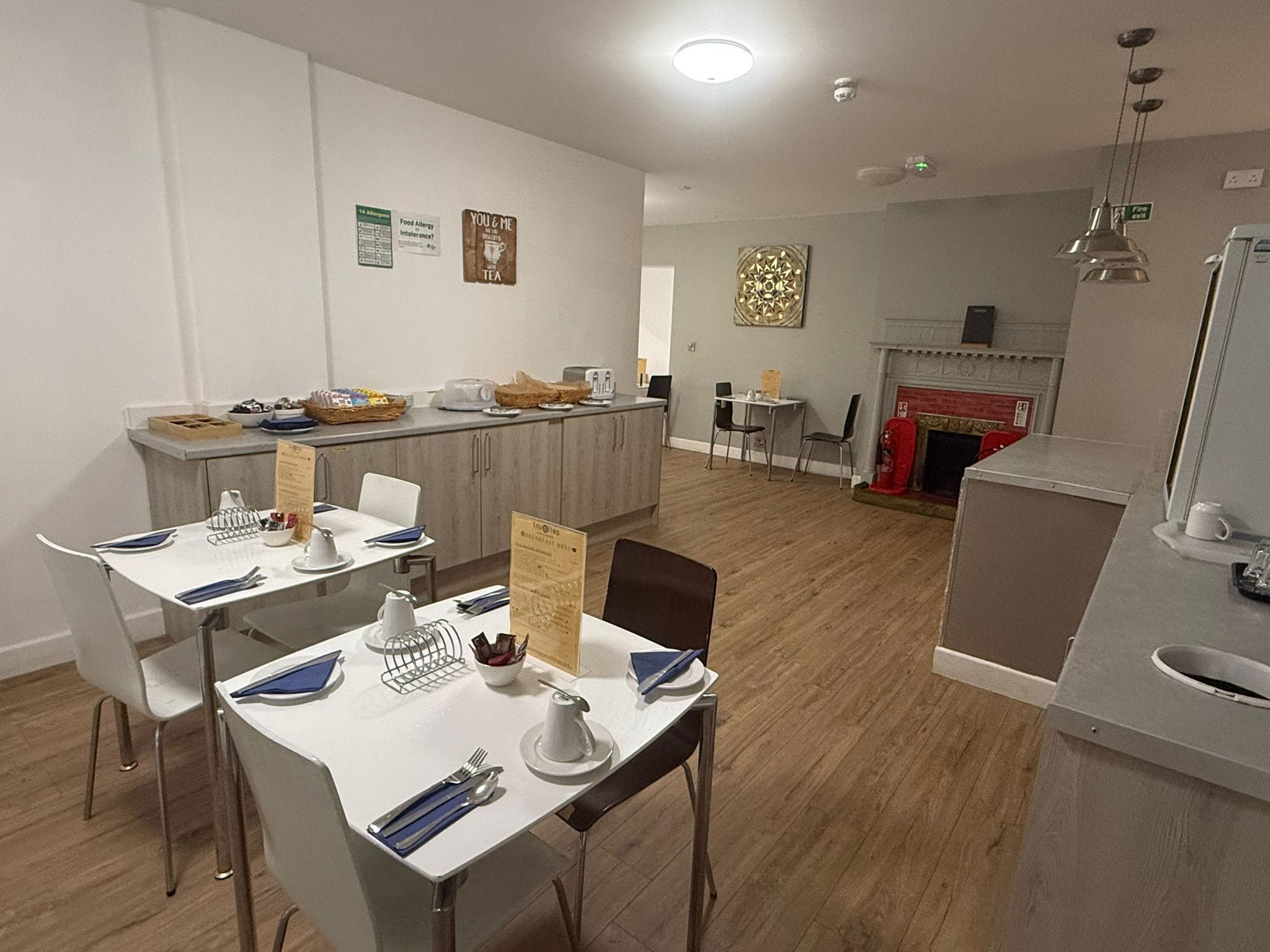 Een moderne eetruimte met witte tafels en stoelen, een keuken op de achtergrond en een decoratief wandkunstwerk.
