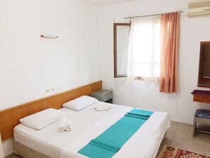 Ein ordentlich bezogenes Bett mit weißen Laken und einer türkisfarbenen Decke, ein kleiner Nachttisch, ein Spiegel und ein Fenster mit Vorhängen.