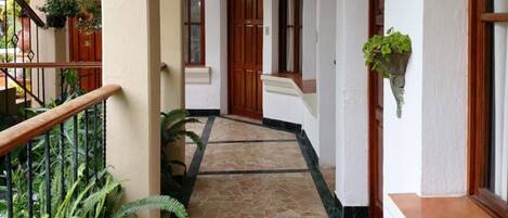 A corridor with a tiled floor, white walls, and wooden doors. There are potted plants along the corridor.
