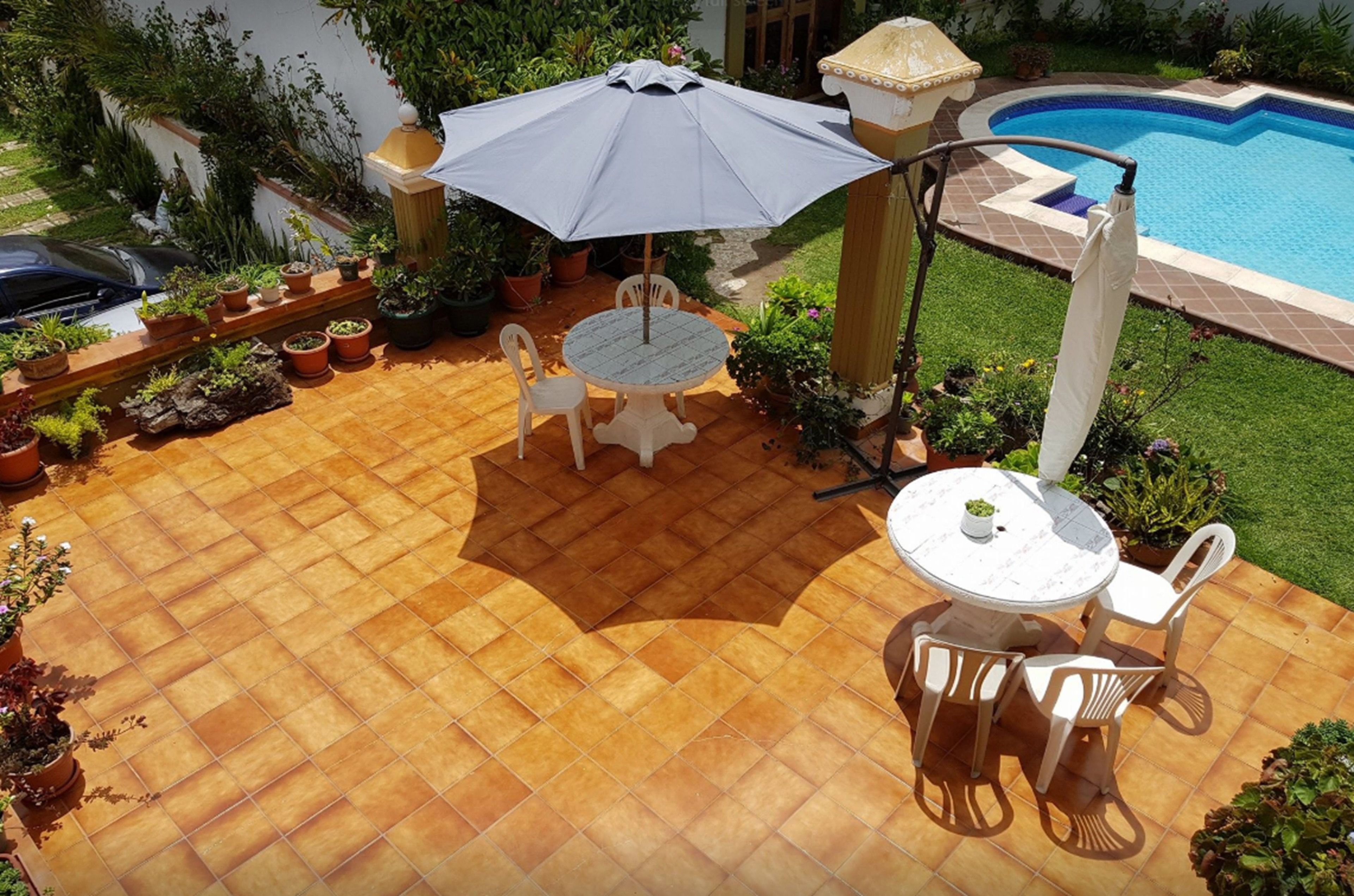 Eine Terrasse mit rundem Tisch und Stühlen, einem großen Sonnenschirm und einem Schwimmbecken im Hintergrund.