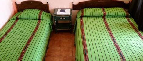 Una habitación con dos camas individuales, cada una con ropa de cama verde a rayas y cabeceros de madera. Hay una mesita de noche pequeña con una lámpara y un libro encima.