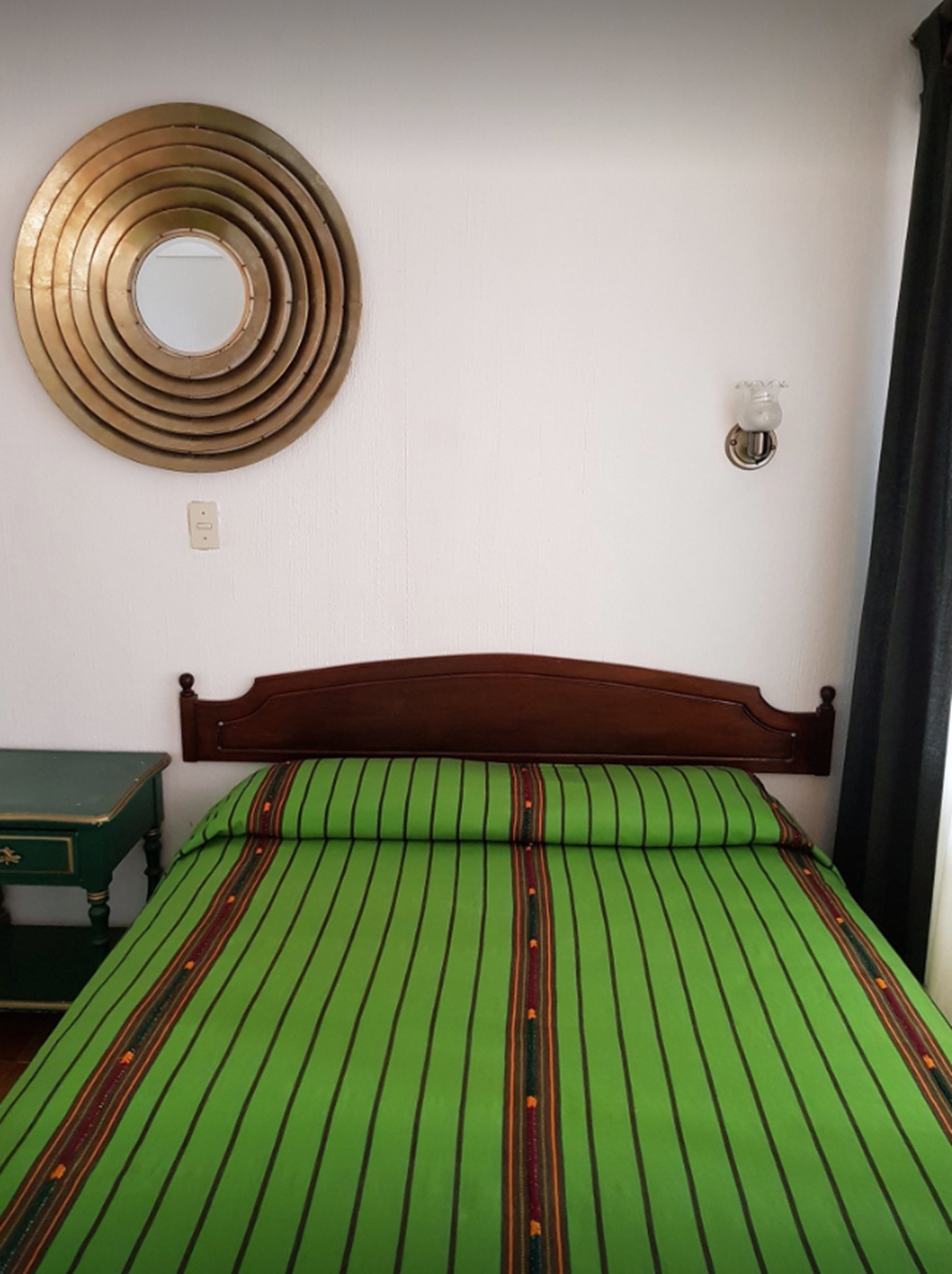 A room with a bed, a wooden headboard, a circular mirror, a bedside table, and a wall-mounted light switch.