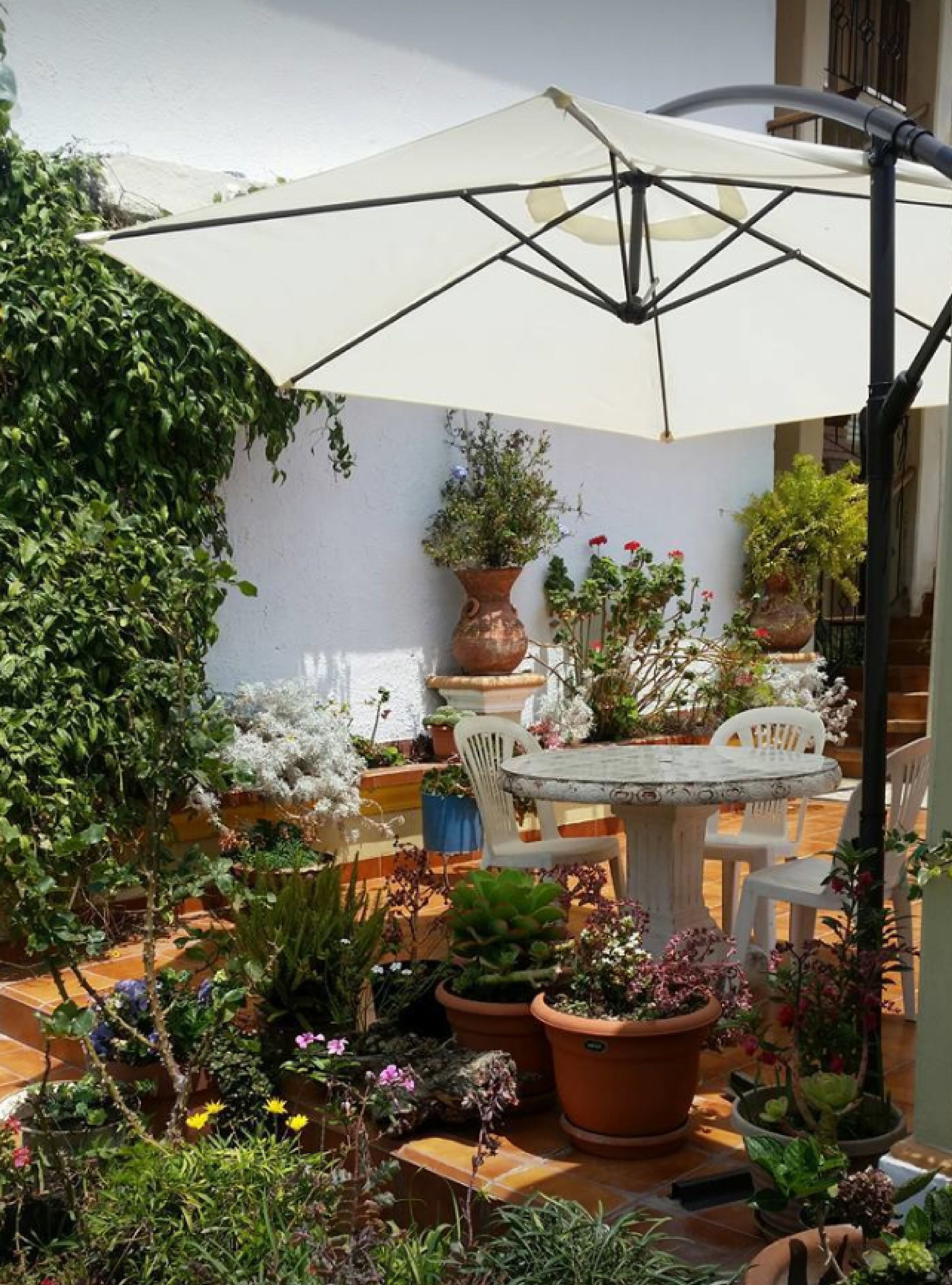 Un patio con mesa y sillas bajo un paraguas blanco, rodeado de macetas con plantas y flores.