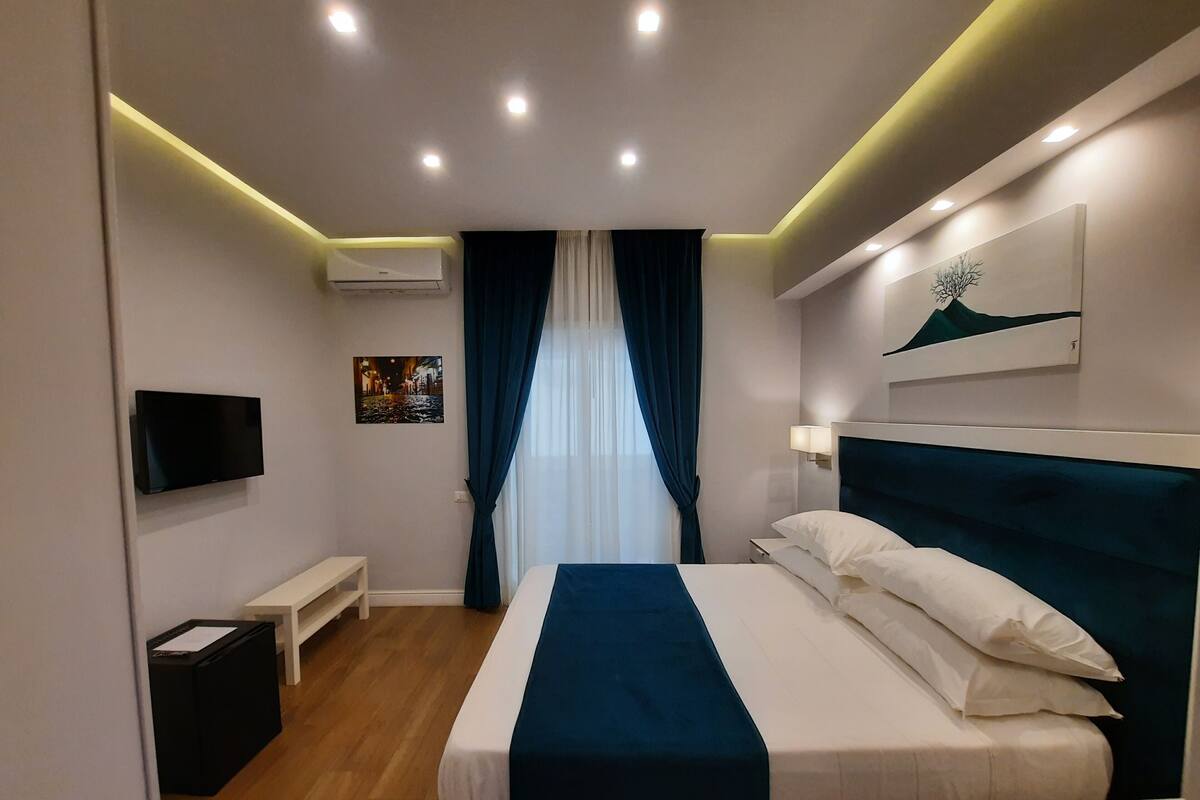 Ein modernes Hotelzimmer mit einem ordentlich bezogenen Bett, einem wandmontierten Flachbildfernseher, einem Nachttisch und einem mit Vorhängen versehenen Fenster.