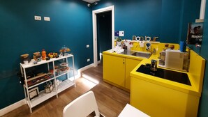 A modern kitchen with teal walls, yellow cabinets, and a white countertop. There is a white chair and a small white table. - Gea Suite (Naples)