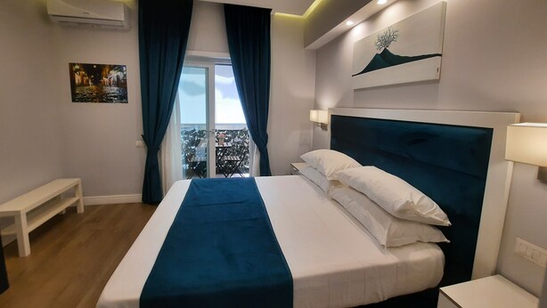A modern hotel room with a large bed, a balcony with a view, a painting on the wall, and an air conditioning unit. - Gea Suite (Naples)