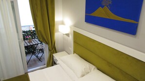 A hotel room with a bed, a painting of a mountain and tree, a window with curtains, and a view of a balcony with plants. - Gea Suite (Naples)