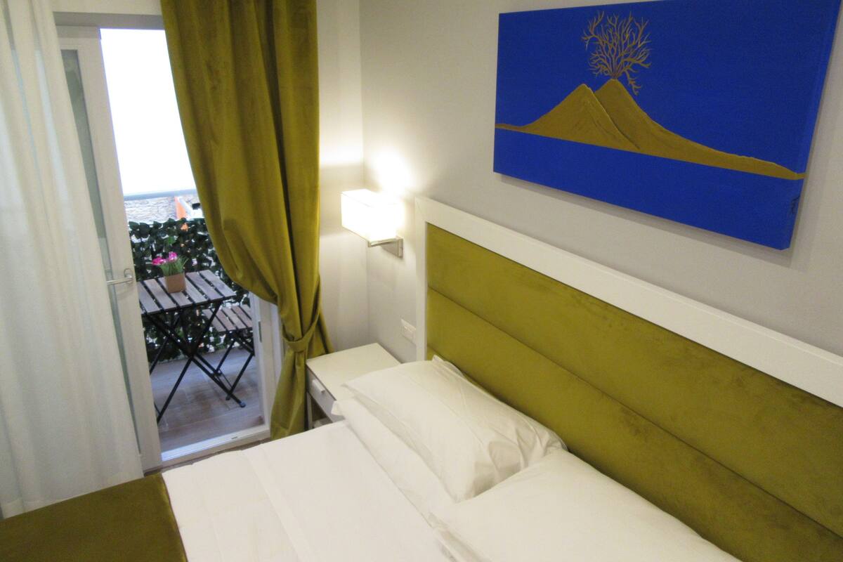 Ein Hotelzimmer mit Bett, einem Bild von Berg und Baum, einem Fenster mit Vorhängen und Blick auf einen Balkon mit Pflanzen.
