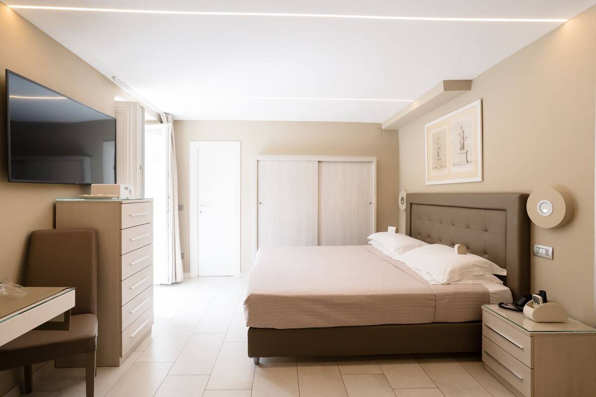 Ein Schlafzimmer mit Bett, Nachttisch, Sessel, Schreibtisch und einem an der Wand montierten Fernseher.