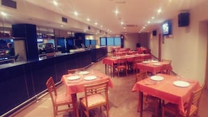 Een restaurant met meerdere tafels en stoelen, een toonbank met een televisiescherm, en een betegelde vloer.