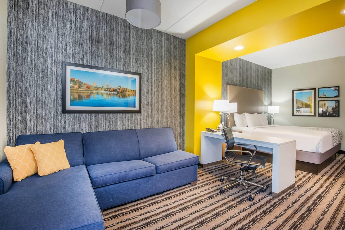 A hotel room with a blue sofa, a desk with a chair, a bed, and a large framed picture on the wall.